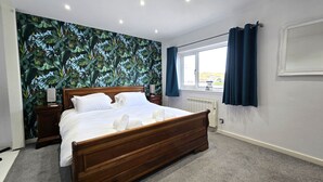 A neatly made bed with white linens and towels, a wooden headboard, and a wall with a tropical leaf pattern. - Spacious apartment, 2 mins walk to the beach! (Perranporth)