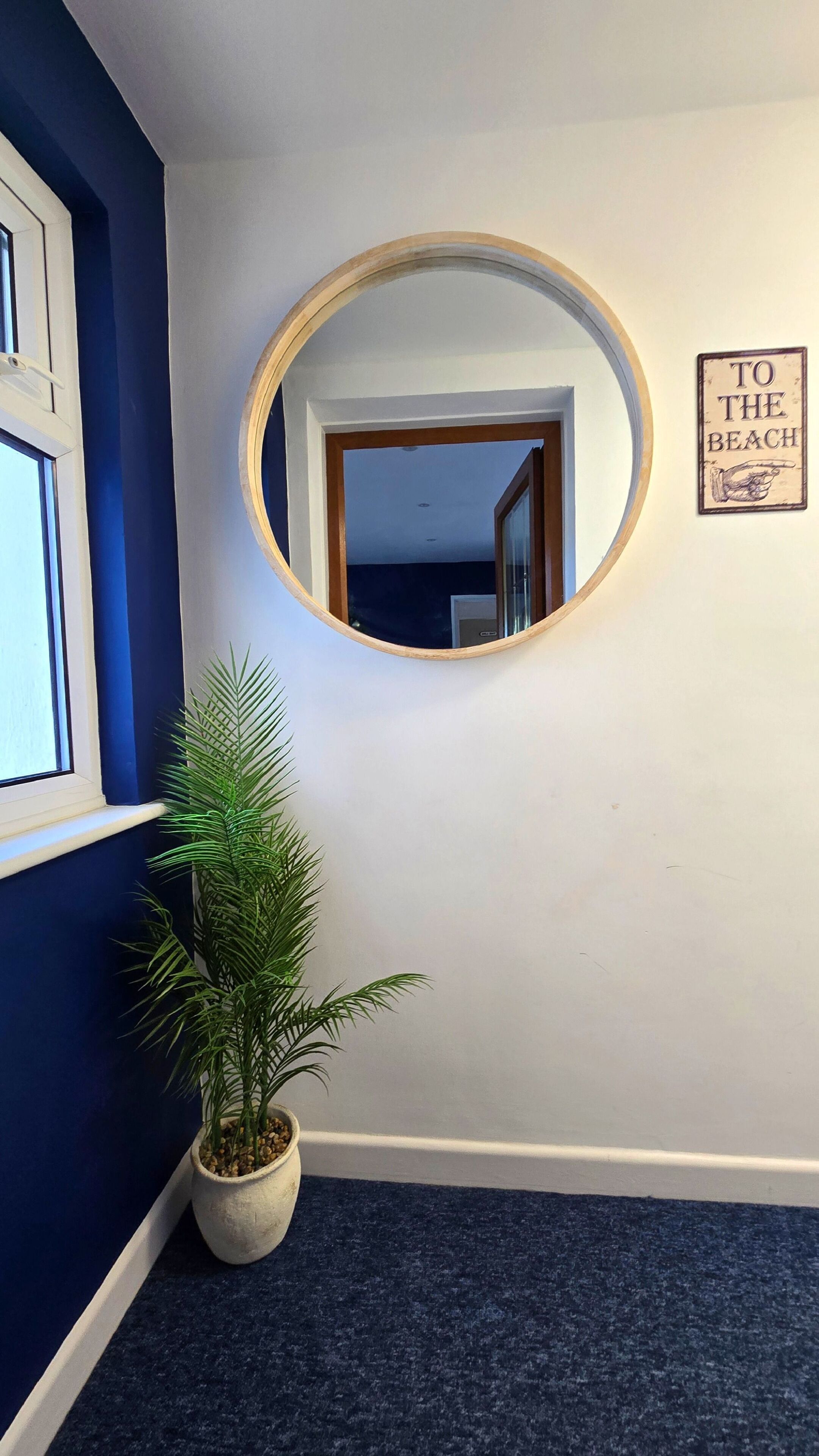 A hallway with a round mirror, a potted plant, and a 'To the Beach' sign.