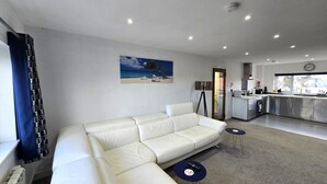 A modern living room with a white sofa, a small round table, and a kitchen area with a sink and stove. - Spacious apartment, 2 mins walk to the beach! (Perranporth)