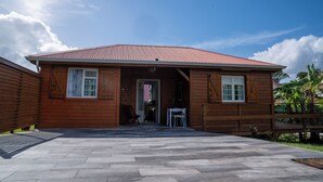 Een houten huis met een betegeld terras, een deur en een klein tafeltje met een stoel.