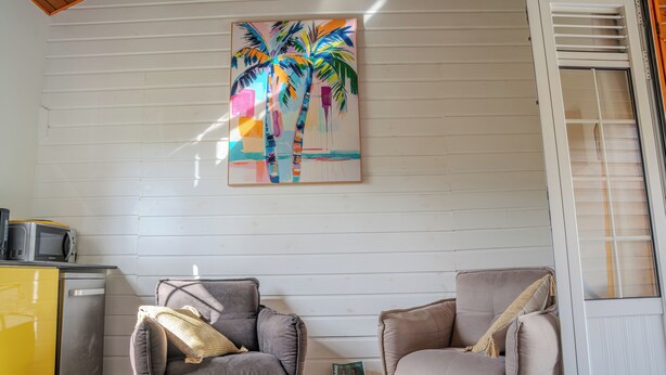 Een kamer met twee fauteuils, een kleine tafel, een schilderij van palmbomen, en een keuken met een magnetron en een geel kastje.