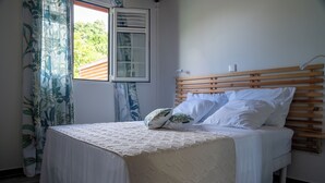 Een net opgemaakt bed met een houten hoofdbord, witte beddengoed en een raam met uitzicht op groen.
