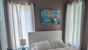A bedroom with a white bed, a small round table with a lamp, a painting on the wall, and a view of the outdoors through a window with white curtains. - Cozy 2-bedroom cottage in charming Council Bluffs with AC (Council Bluffs)