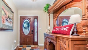 Una puerta principal de madera con un panel de vidrio, un cojín rojo con el texto "WELCOME HOME" y una lámpara sobre una consola de madera.