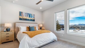 A bedroom with a large bed, two bedside tables, a ceiling fan, a painting, and a view of a snowy landscape through a large window. - Rendezvous 4BR Retreat | New Hot Tub, Mtn Views (Winter Park)