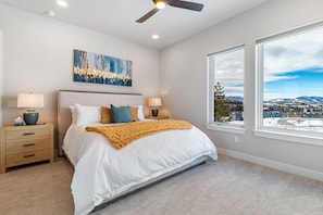 A bedroom with a large bed, two bedside tables, a ceiling fan, a painting, and a view of a snowy landscape through a large window. - Rendezvous 4BR Retreat | New Hot Tub, Mtn Views (Winter Park)