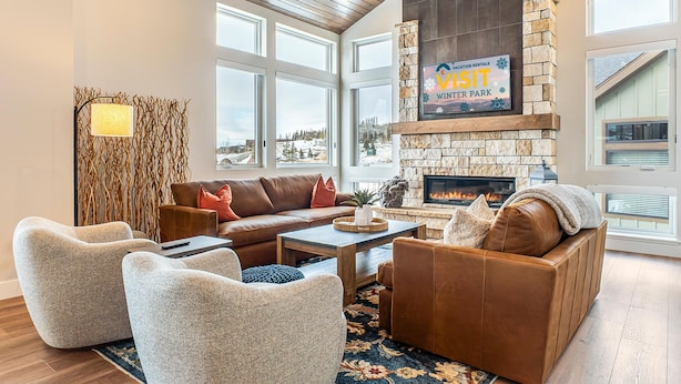 A cozy living room with a stone fireplace, leather sofas, and a view of a snowy landscape through large windows. - Rendezvous 4BR Retreat | New Hot Tub, Mtn Views (Winter Park)