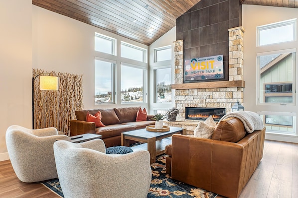 A cozy living room with a stone fireplace, leather sofas, and a view of a snowy landscape through large windows. - Rendezvous 4BR Retreat | New Hot Tub, Mtn Views (Winter Park)
