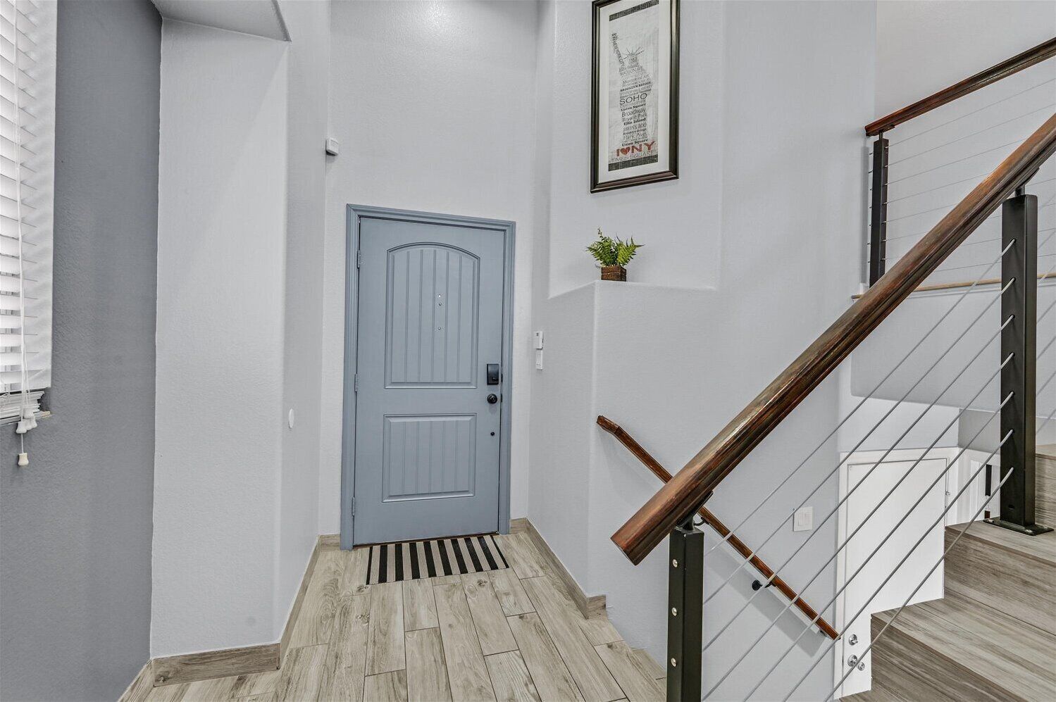 Um corredor com uma porta azul, uma escada com corrimãos de madeira e um pequeno vaso com planta na parede.