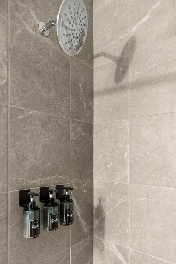 Une douche équipée d’un pommeau de douche à effet pluie et de trois distributeurs muraux contenant du shampoing, de l’après-shampoing et du gel douche.