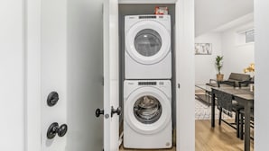 A compact laundry room with two stacked Whirlpool front-loading washing machines, a dining area with a table and chairs, and a potted plant. - CozyWolf (New 2 Bedroom Suite) (Calgary)