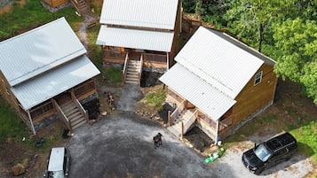 A wooden cabin complex with multiple buildings, a central fire pit, and two vehicles parked in a gravel area.
