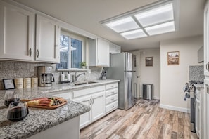 A modern kitchen with white cabinets, granite countertops, a stainless steel refrigerator, and a window with a view of trees. - Kaweah Oaks Cabin: Firepit, Hot Tub, Game Room (Three Rivers)