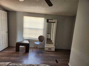 Een kamer met een houten vloer, een bankje, een stoel, een grote spiegel en een raam met jaloezieën.