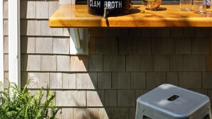 Un espace bar extérieur en bois, avec une enseigne indiquant « CLAM BROTH », des bouteilles et un saladier rempli de fruits.