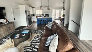 Amplia sala de estar con un sofá de cuero marrón, una mesa de centro y una alfombra estampada. Se ve una cocina al fondo, con armarios blancos y una isla azul.