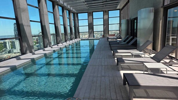 Et rooftop-infinity pool med krystalklart, blåt vand, omgivet af liggestole og store vinduer med udsigt over byen.