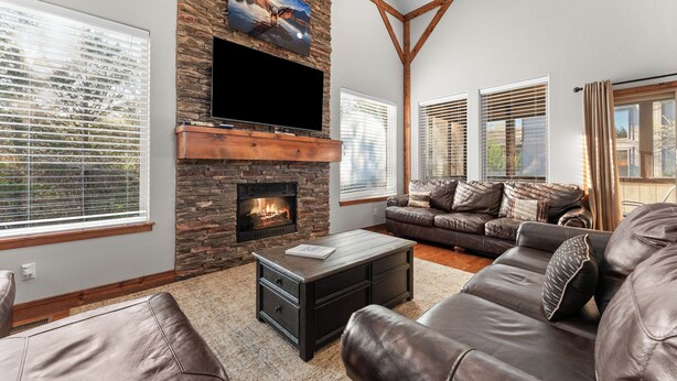 Una sala de estar con chimenea de piedra, sofás de cuero, televisor de pantalla plana y ventanas grandes con persianas.