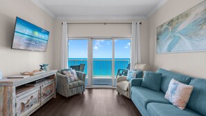 A living room with a view of the ocean, a TV on the wall, and a sofa. - 18th Floor!  Indoor/Outdoor Pools. Free Daily Activities + Beach Service (Gulf Shores)