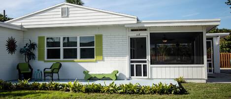 A white house with a covered porch, two chairs, a bench, and a sign that reads "BEACH HOUSE".