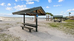 Ein Strand mit einer hölzernen Bank, einem Sonnensegel und einer Rollstuhlbereitheitstafel, sowie Palmen.