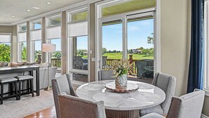 Ein geräumiges Zimmer mit großem Esstisch, bequemen Sitzmöglichkeiten und großen Fenstern mit Blick auf einen Golfplatz.