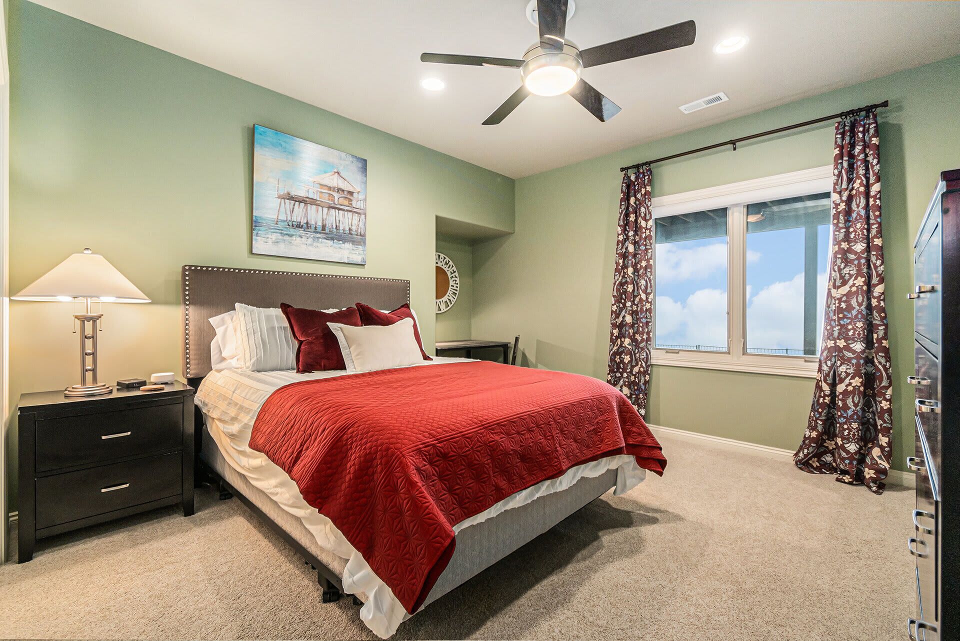 Une chambre à coucher avec un lit, des tables de chevet, un ventilateur de plafond, une fenêtre avec des rideaux et un tableau au mur.