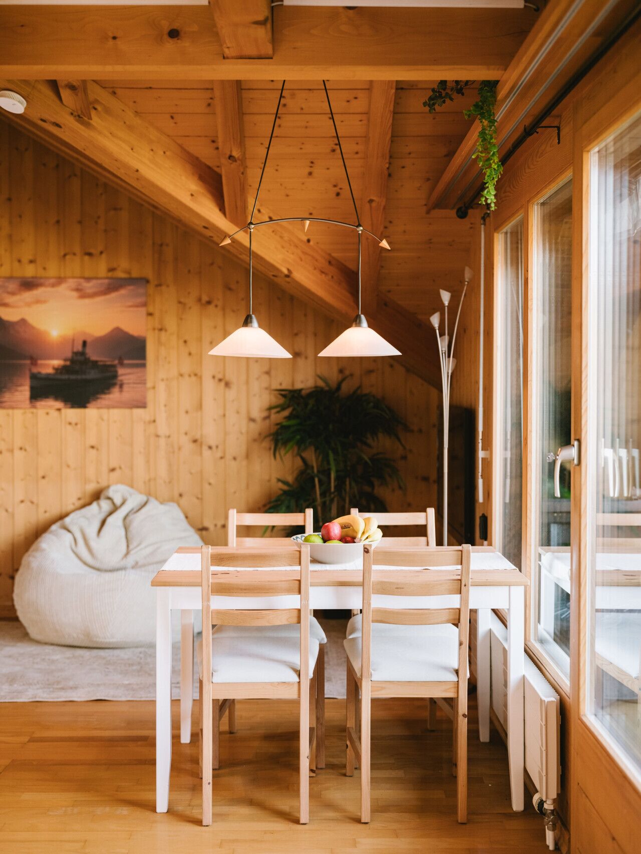 A wooden interior with a dining table, chairs, a bean bag, and a large window.