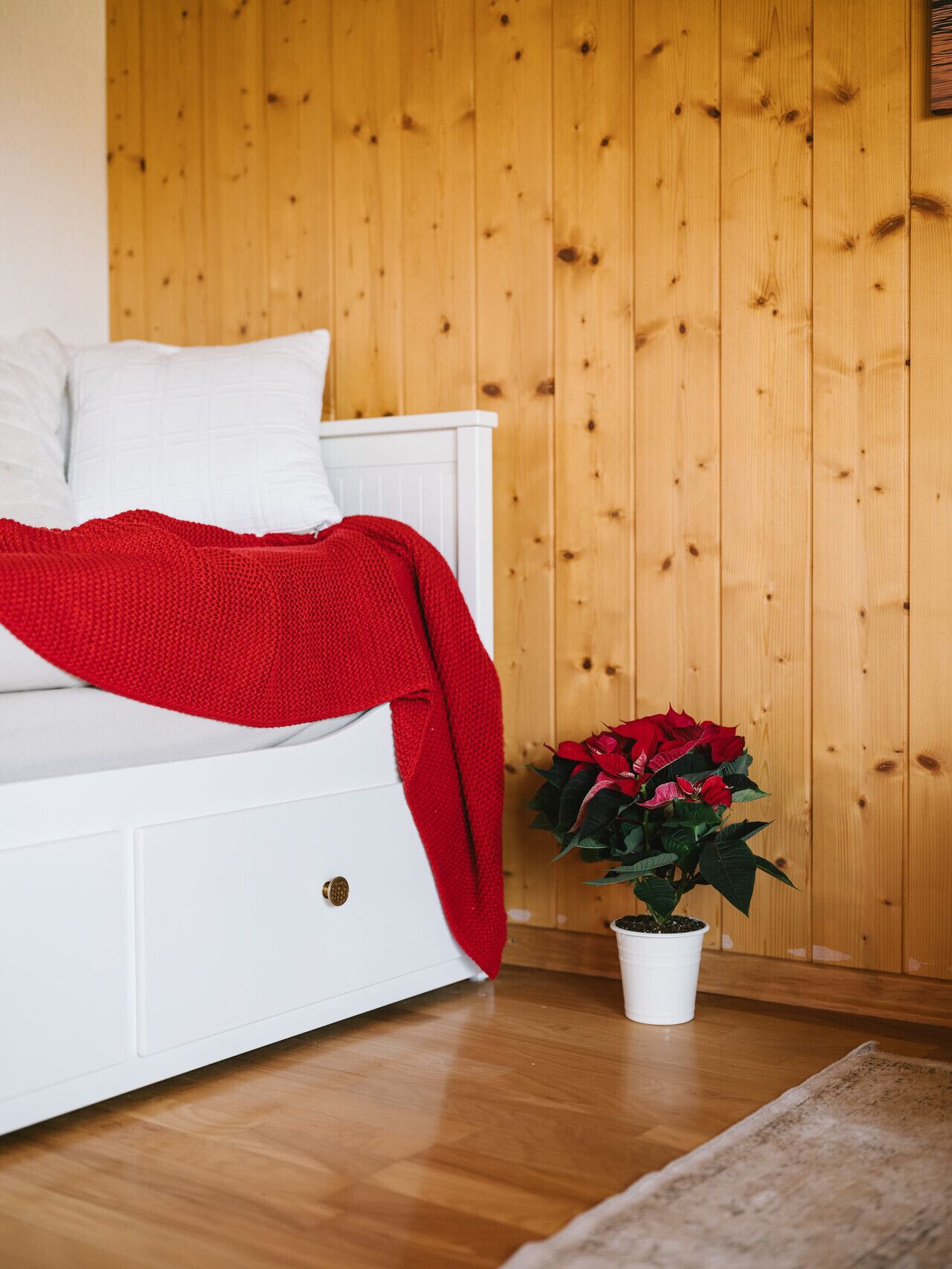 Ein weißes Bett mit roter Decke, einem weißen Kopfkissen und einem Blumentopf mit roten Blüten.