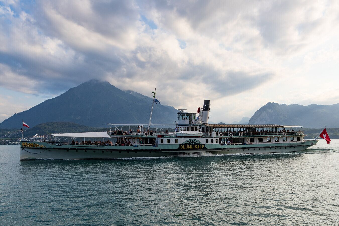 A boat with the name "BLÜMLISALP" on its side, sailing on a body of water with mountains in the background.
