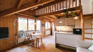 Un espacio amplio con piso de madera, una zona de comedor con mesa y sillas, una cocina con gabinetes blancos y microondas, y ventanas grandes que ofrecen vistas al exterior.