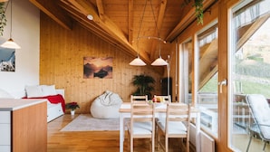 Interior acogedor de una cabaña con paredes de madera, una zona de comedor con mesa y sillas, un sillón de peluche, y un ventanal con vistas al exterior.