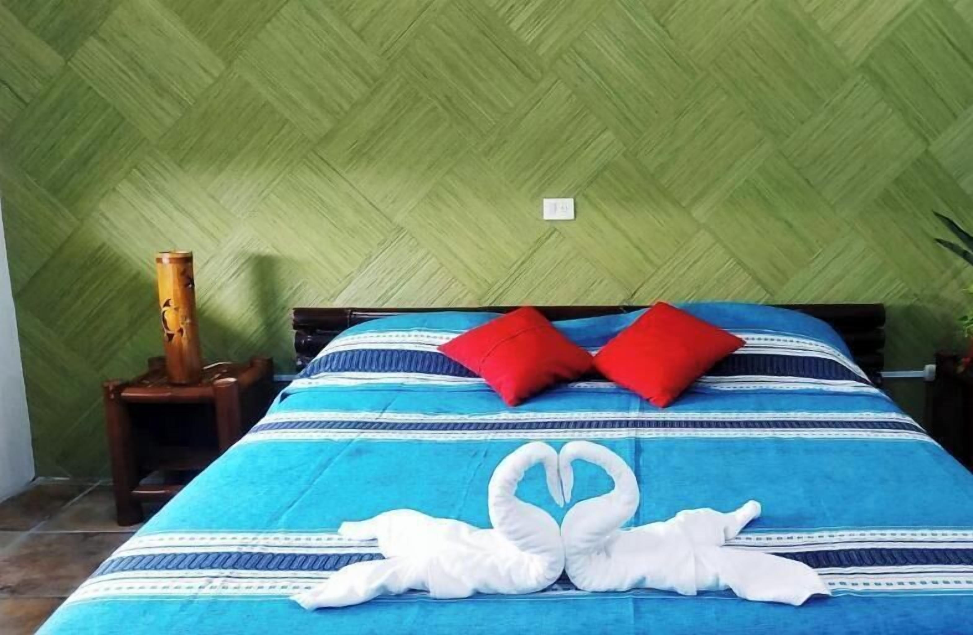 Una cama con una colcha azul y blanca a rayas, dos almohadas rojas y una toalla blanca dispuestas en forma de corazón sobre la cama.