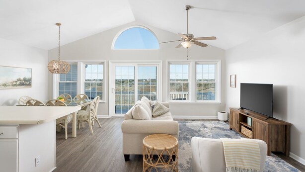 A spacious living area with a high ceiling, large windows, a ceiling fan, a television, and a dining area with wicker furniture.