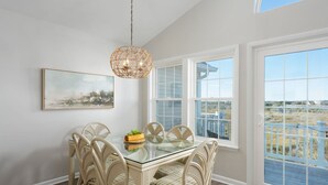 A dining area with a glass table, wicker chairs, a hanging light fixture, and a view of the beach through large windows.