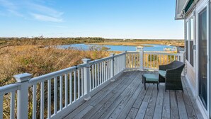 A wooden deck with white railings overlooking a marshy landscape and a body of water.