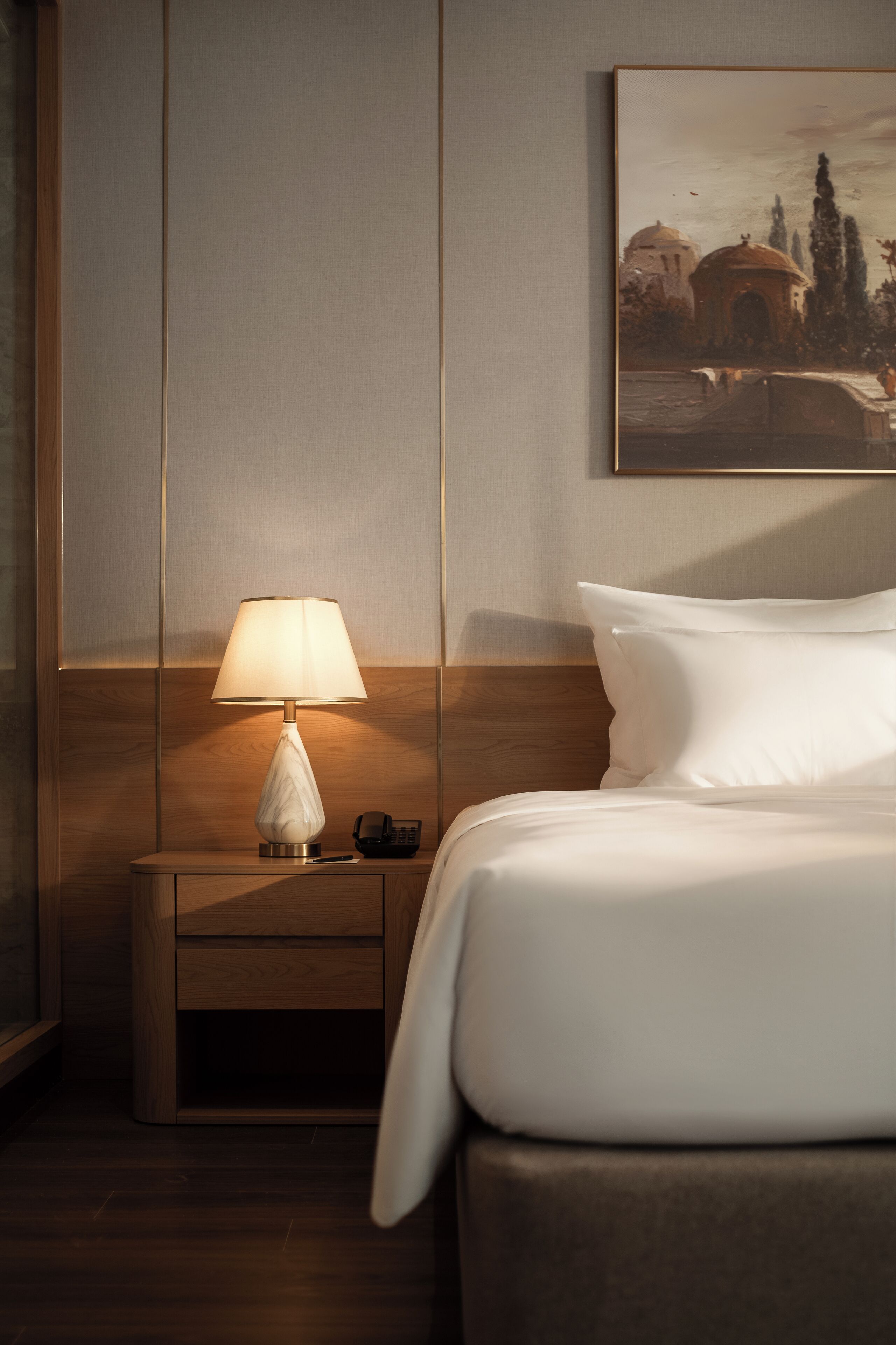 A hotel room with a wooden nightstand, a white lamp, a bed with white bedding, and a framed painting on the wall.