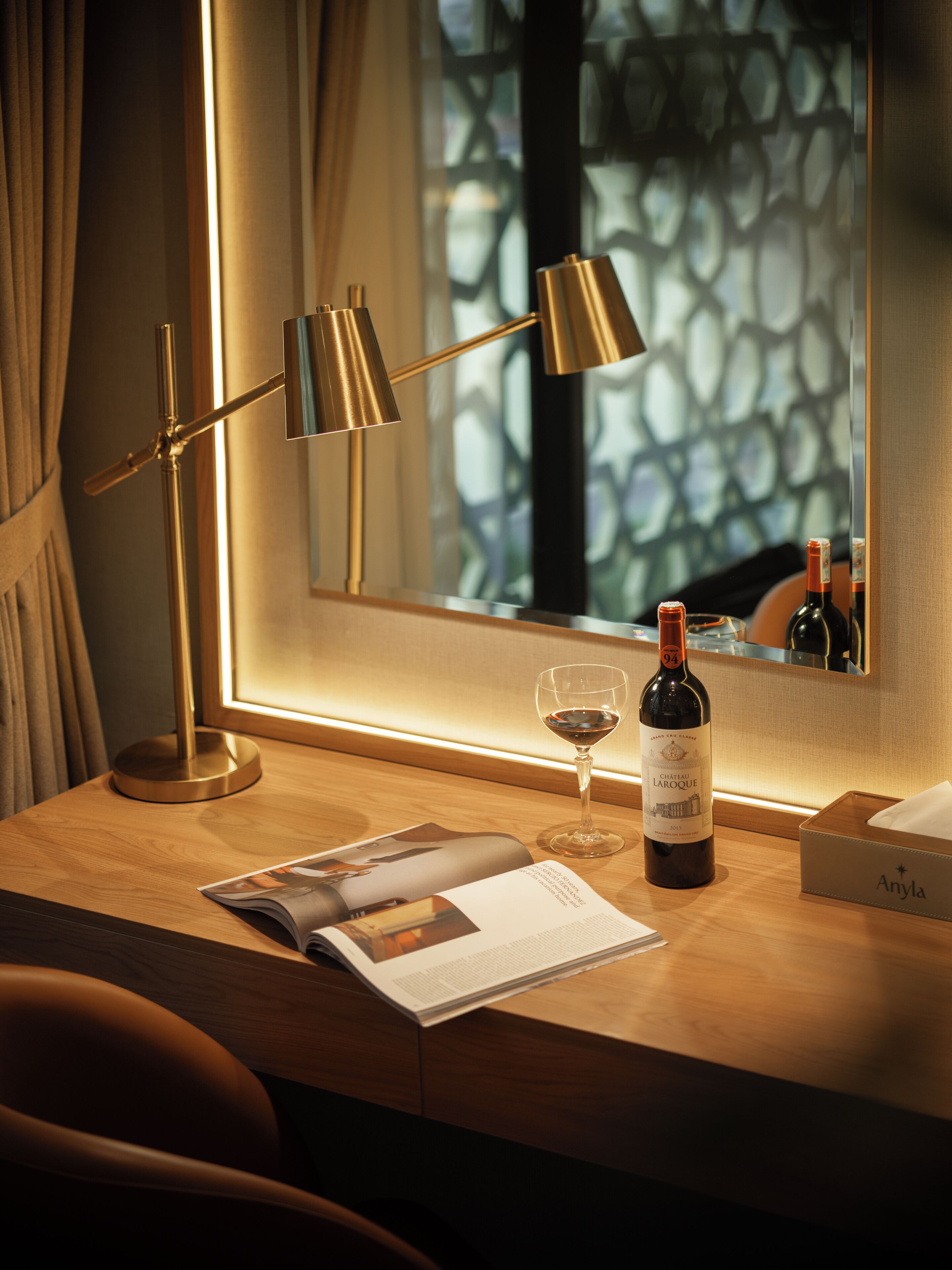 Un bureau en bois avec un magazine, une bouteille de vin, un verre et une lampe.