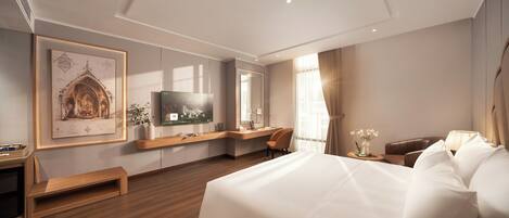 A hotel room with a large bed, a desk with a flat-screen TV, a seating area with a chair, and a framed architectural drawing on the wall.