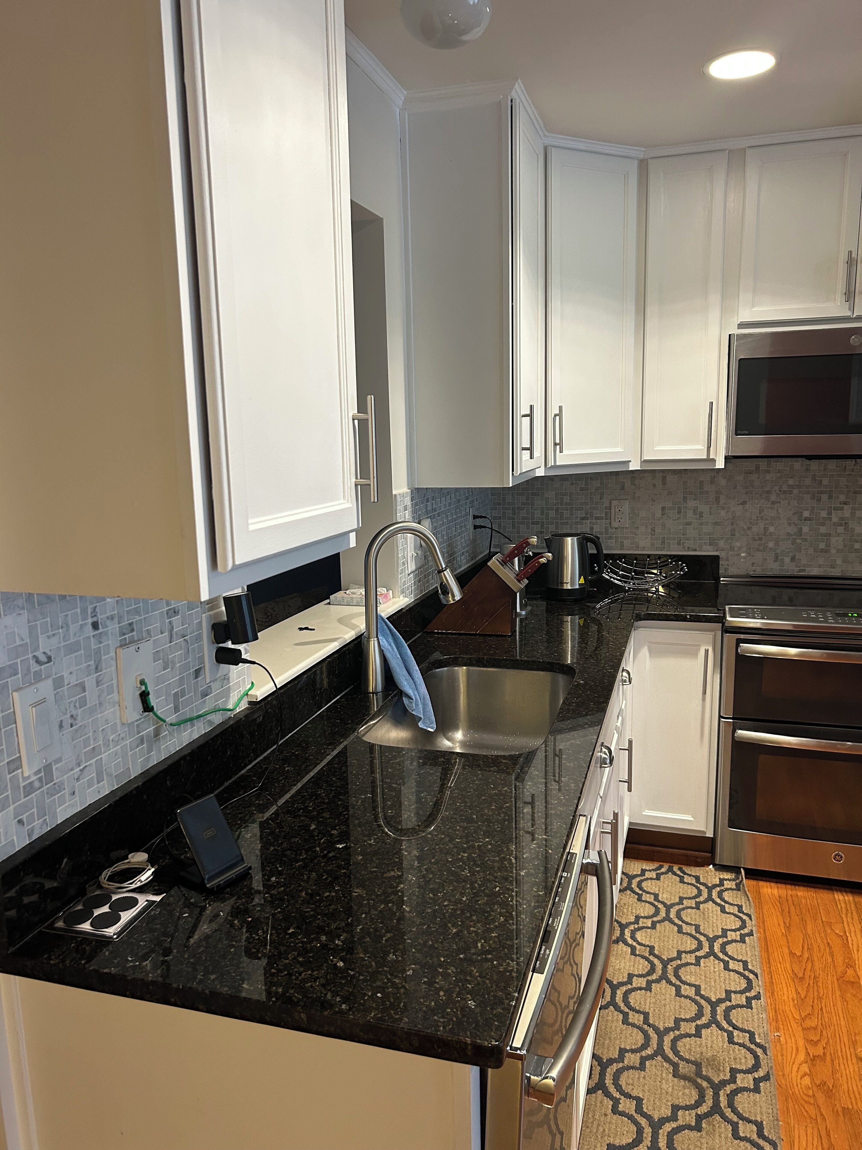 A modern kitchen with white cabinets, a black granite countertop, stainless steel appliances, and a patterned rug.