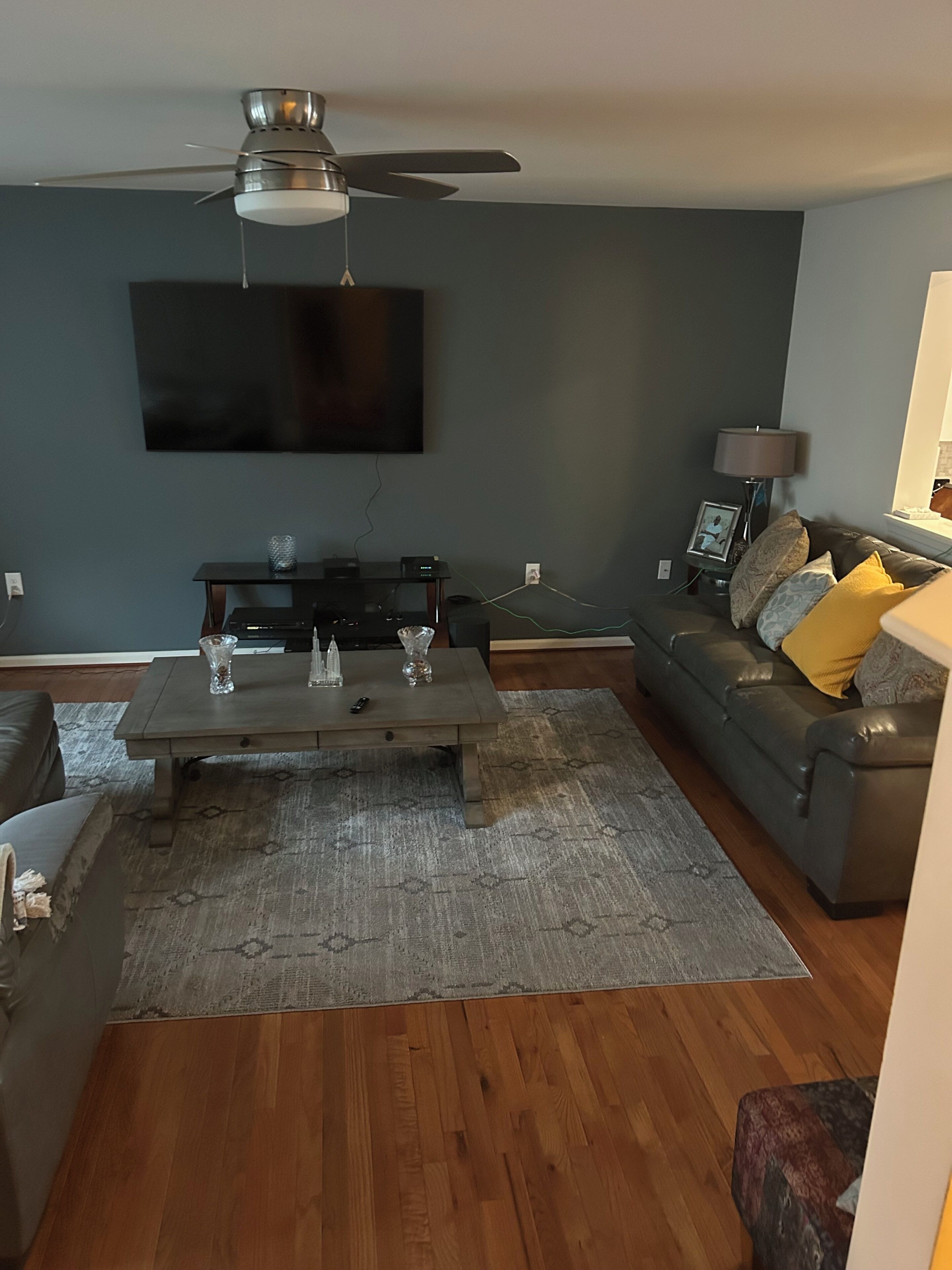 A living room with a ceiling fan, a flat-screen TV mounted on the wall, a dark-colored sofa with yellow cushions, and a wooden coffee table with decorative items.