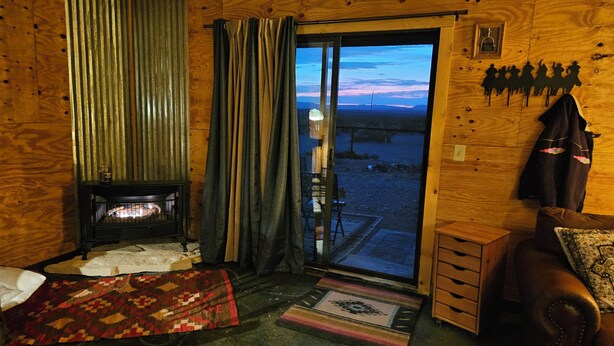 A cozy cabin interior with a wood-burning stove, a sliding glass door leading to a snowy outdoor area, and a leather couch. - THE FAR SIDE OF THE MOON
Off Grid 1-bedroom  "Cowboy Camp" in the desert (Terlingua)