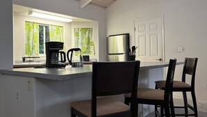 Eine Küche mit Kaffeemaschine, Wasserkocher und Kühlschrank. Zwei Stühle und ein Fenster mit Vorhängen.