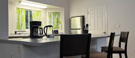A kitchen with a coffee maker, kettle, and refrigerator. There are two chairs and a window with curtains.