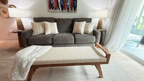 Una sala de estar moderna con un sofá gris, cojines estampados, una manta blanca y una mesa de centro de madera. Una obra de arte colorida cuelga sobre el sofá, y hay dos lámparas sobre mesitas auxiliares.