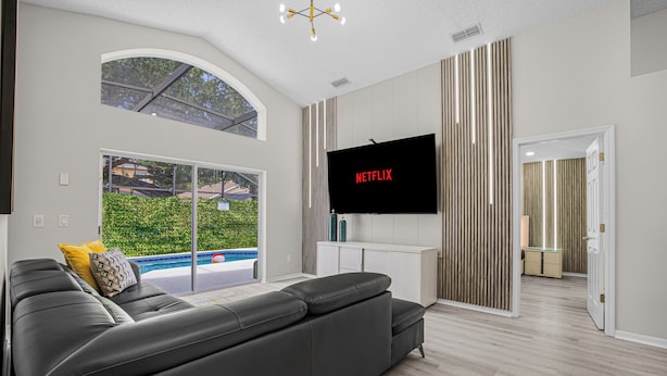 Een moderne woonkamer met een grote flatscreen-tv, een grijze bank en uitzicht op een zwembad door grote ramen.