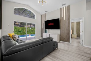 Ett modernt vardagsrum med en stor platt-TV, en grå soffa och utsikt över en swimmingpool genom stora fönster.