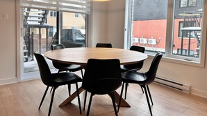 Un espace repas avec une table ronde en bois et des chaises noires, de grandes fenêtres donnant sur un bâtiment et du mobilier d’extérieur, ainsi qu’un luminaire suspendu au plafond.