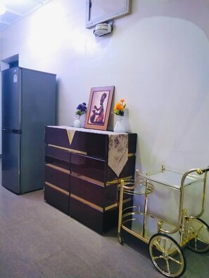 Une pièce avec un réfrigérateur noir, un meuble en bois foncé doté de tiroirs, une petite table ornée d’un tableau et de fleurs, et un porte-vélo avec un cadre doré.