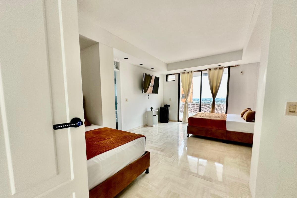 Habitación de hotel con cama, escritorio, silla, televisor y balcón con cortinas.